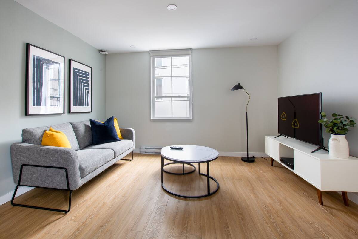 Un salon moderne avec un canapé gris, une table basse ronde, un lampadaire et un téléviseur posé sur un meuble blanc.