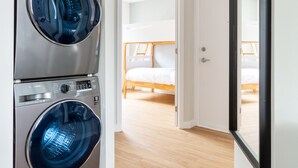 A laundry room with two stacked front-loading washers and dryers, a wooden floor, and a door leading to a bedroom with a bed.