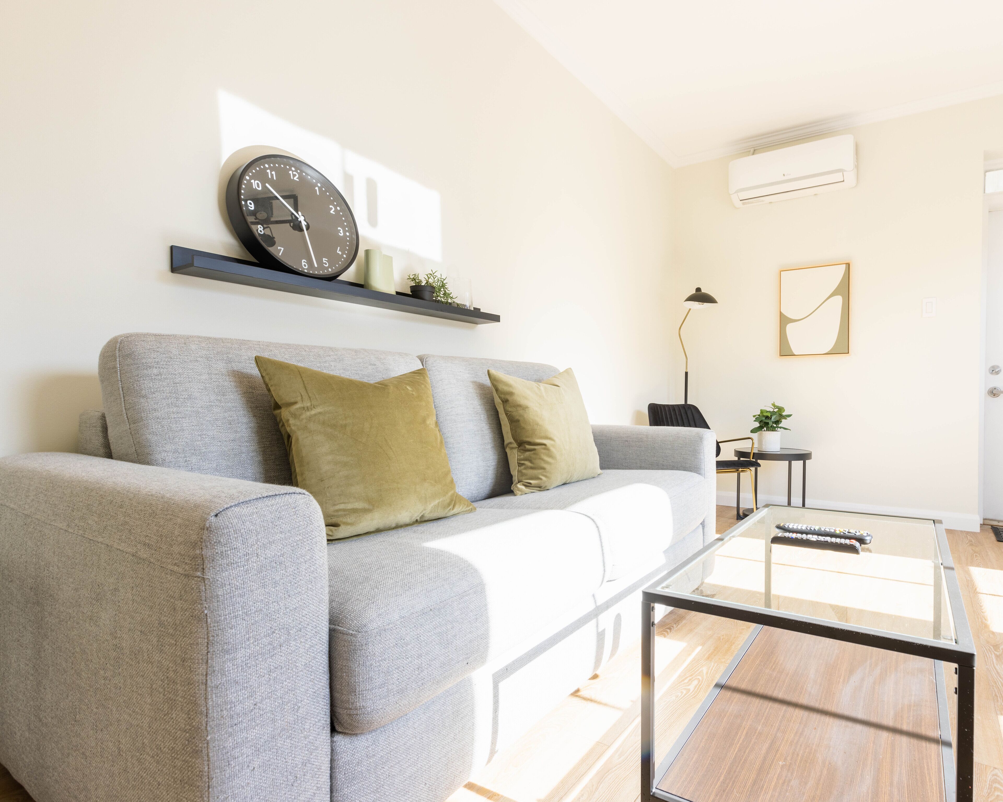 En moderne stue med en grå sofa, to puder, et glasbord, et ur, et lille sengebord med en lampe og en vægmonteret klimaanlæg.