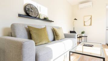 En moderne stue med en grå sofa, to puder, et glasbord, et ur, et lille sengebord med en lampe og en vægmonteret klimaanlæg.