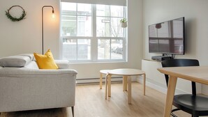 A modern living room with a grey sofa, yellow cushions, a round table, a TV, and a large window with a view of the street.