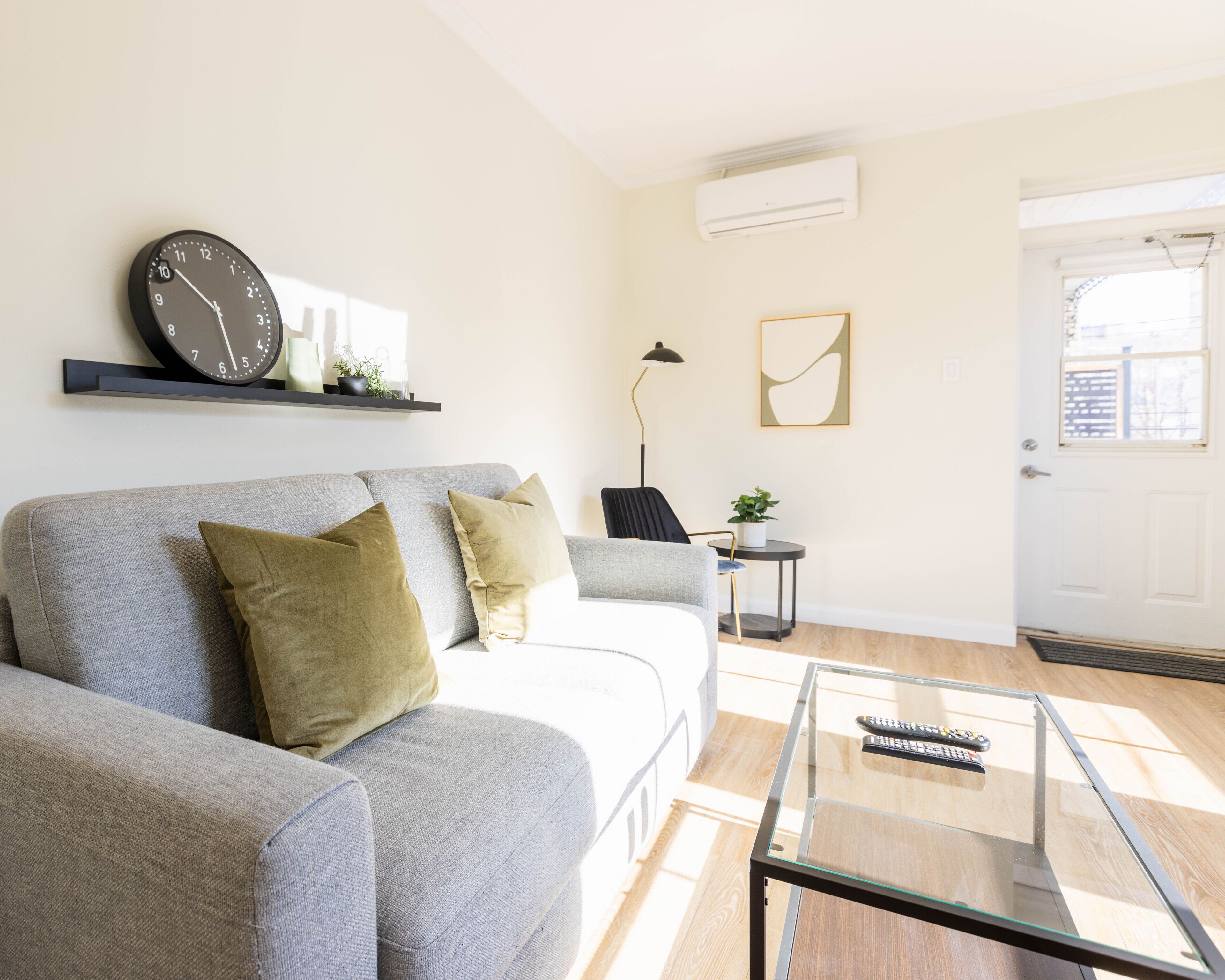 A modern living room with a grey sofa, a glass coffee table, a black clock, and a door with a window view.