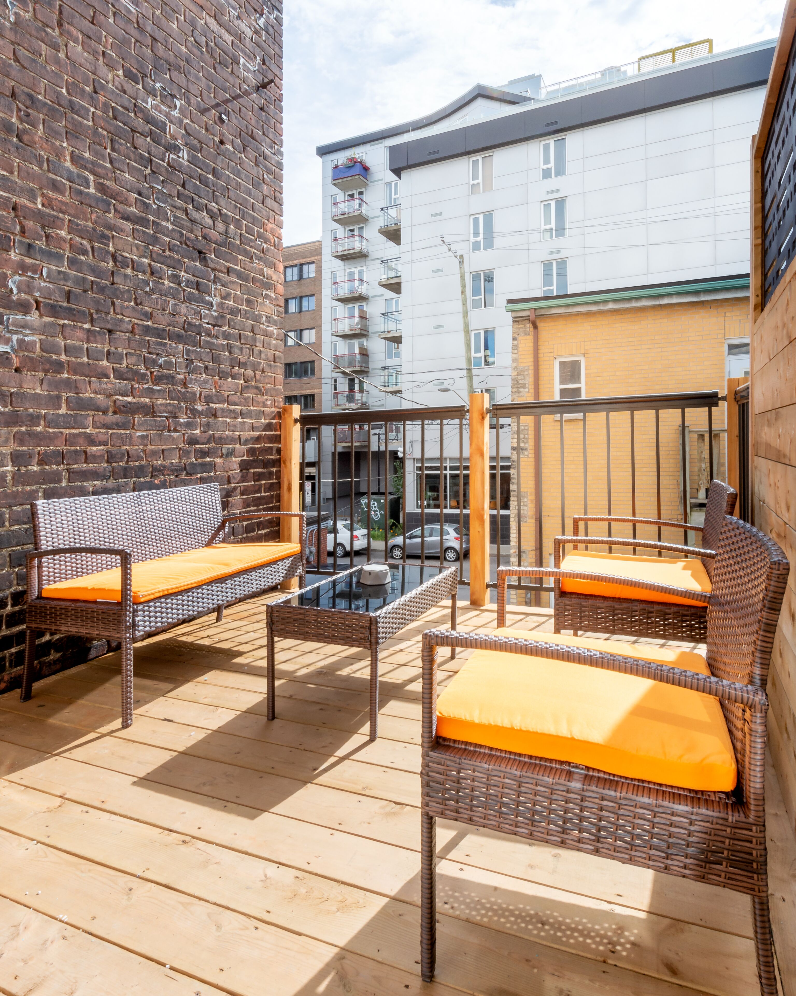 An outdoor seating area with wicker furniture and orange cushions, a brick wall, and a view of a multi-story building in the background.