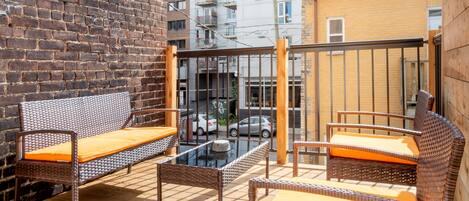 An outdoor seating area with wicker furniture and orange cushions, a brick wall, and a view of a multi-story building in the background.
