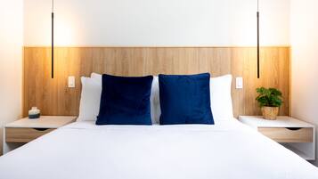 Una cama bien tendida con sábanas blancas y dos almohadas azul oscuro, flanqueada por mesitas de noche de madera con cajones blancos y un portalápices tejido.