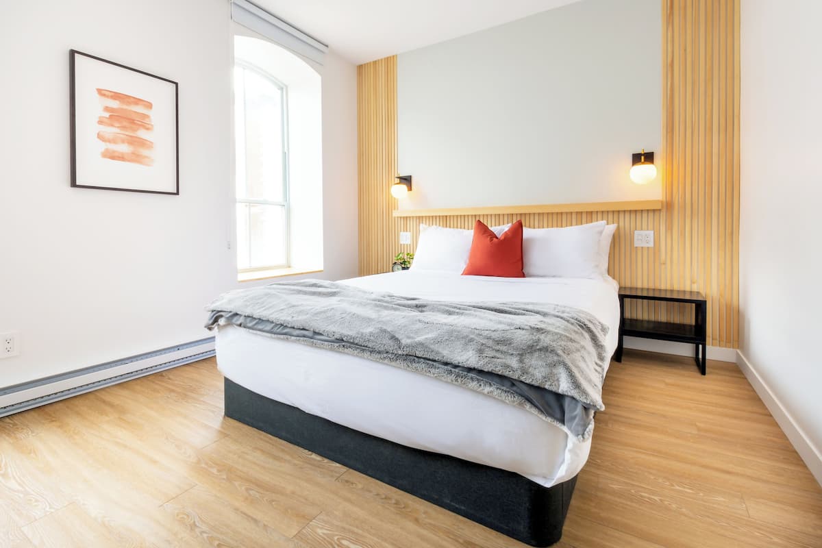 Une chambre moderne avec un grand lit, un plancher en bois, un luminaire fixé au mur et une œuvre d’art encadrée.