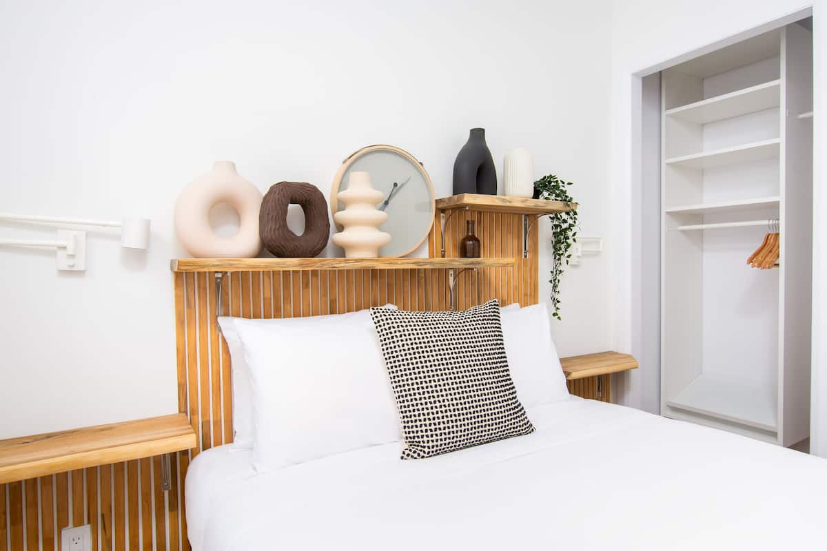 Une chambre à coucher bien aménagée, avec une tête de lit en lattes de bois, un lit blanc agrémenté d’oreillers à motifs et divers objets décoratifs disposés sur des étagères.