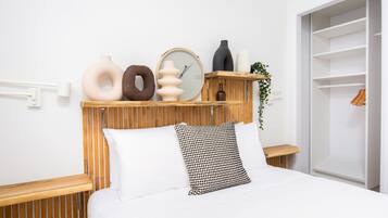 A neatly arranged bedroom with a wooden slat headboard, a white bed with patterned pillows, and various decorative items on shelves.