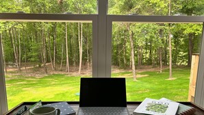 A laptop on a desk with a view of a grassy yard and trees through a window.