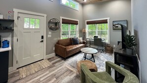 A spacious living room with a brown leather sofa, a round coffee table, and a green armchair. There is a ceiling fan, a TV mounted on the wall, and a window with a view of trees.