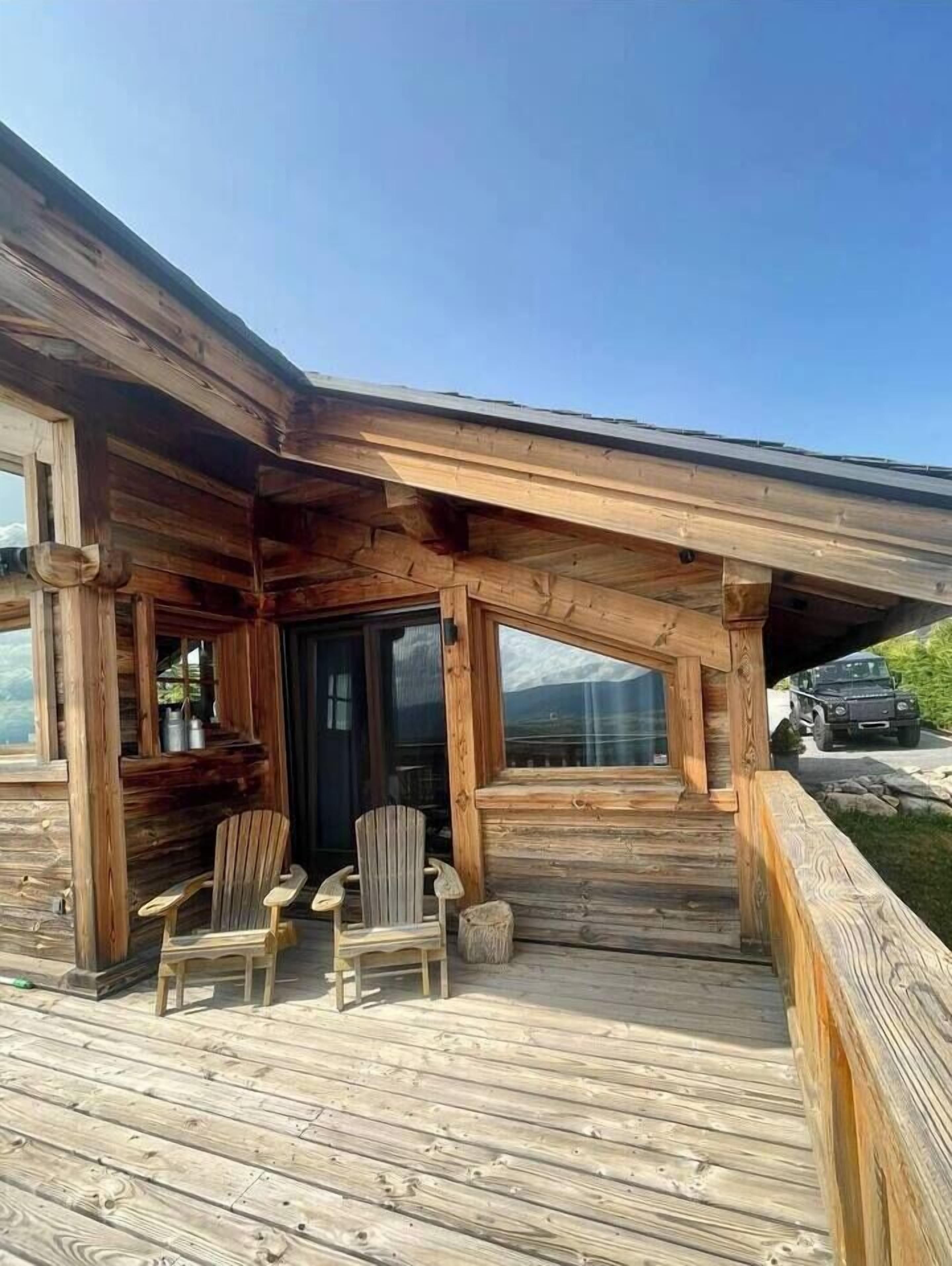 A wooden cabin with a covered porch, two Adirondack chairs, and a vehicle parked outside.