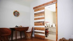 A room with a wooden floor, a chessboard, a chair, a table, a bed with pillows, and a wooden door. - Dos Pinos Riverside Cottage near DT Kerrville (Kerrville)
