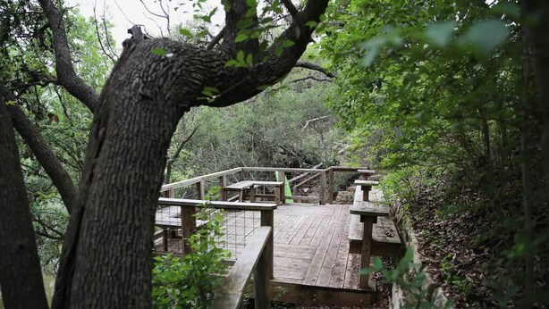 A wooden bridge leading to a wooden deck with benches and picnic tables, surrounded by trees. - Dos Pinos Riverside Cottage near DT Kerrville (Kerrville)