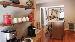 A kitchen with wooden shelves holding mugs and red containers, a coffee maker, and a trash can. - Dos Pinos Riverside Cottage near DT Kerrville (Kerrville)