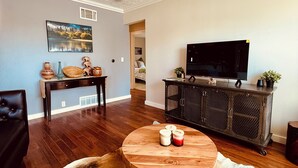 A living room with a round wooden coffee table, a black leather sofa, a television on a black cabinet, and a painting on the wall.
