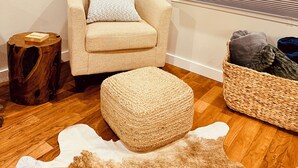 A cozy living room with a beige armchair, a woven ottoman, a wooden side table, and a woven basket with blankets.