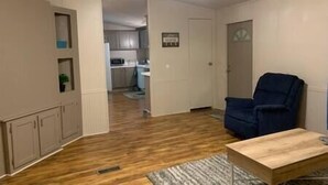 Una sala de estar con un sillón azul, una mesa de centro de madera y una cocina al fondo.