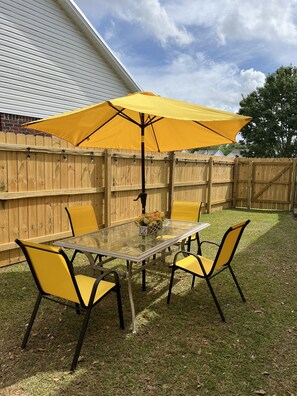 A glass-top table with four yellow chairs and a large yellow umbrella in a backyard with a wooden fence. - Cozy and relaxing beach house, Our Little Pearl in Foley. (Foley)