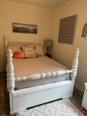 A bedroom with a white four-poster bed, a beige wall, a framed inspirational quote, and a small table with a lamp.