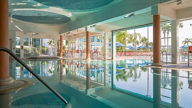 Una piscina interior con paredes de vidrio transparente, una zona junto a la piscina con sombrillas y vistas a palmeras afuera.