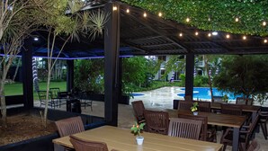 Eine Terrasse im Freien mit Holztischen und -stühlen, Lichterketten und einem Pool im Hintergrund.