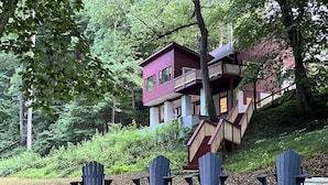 Une cabane à l’extérieur rouge, dotée d’un balcon et d’un escalier menant à celui-ci, entourée d’arbres et d’une pelouse où se trouvent quatre chaises Adirondack.