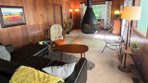 Un salon chaleureux avec un canapé en cuir noir, un plafond en bois, une cheminée noire et un coin repas comprenant une table et des chaises.