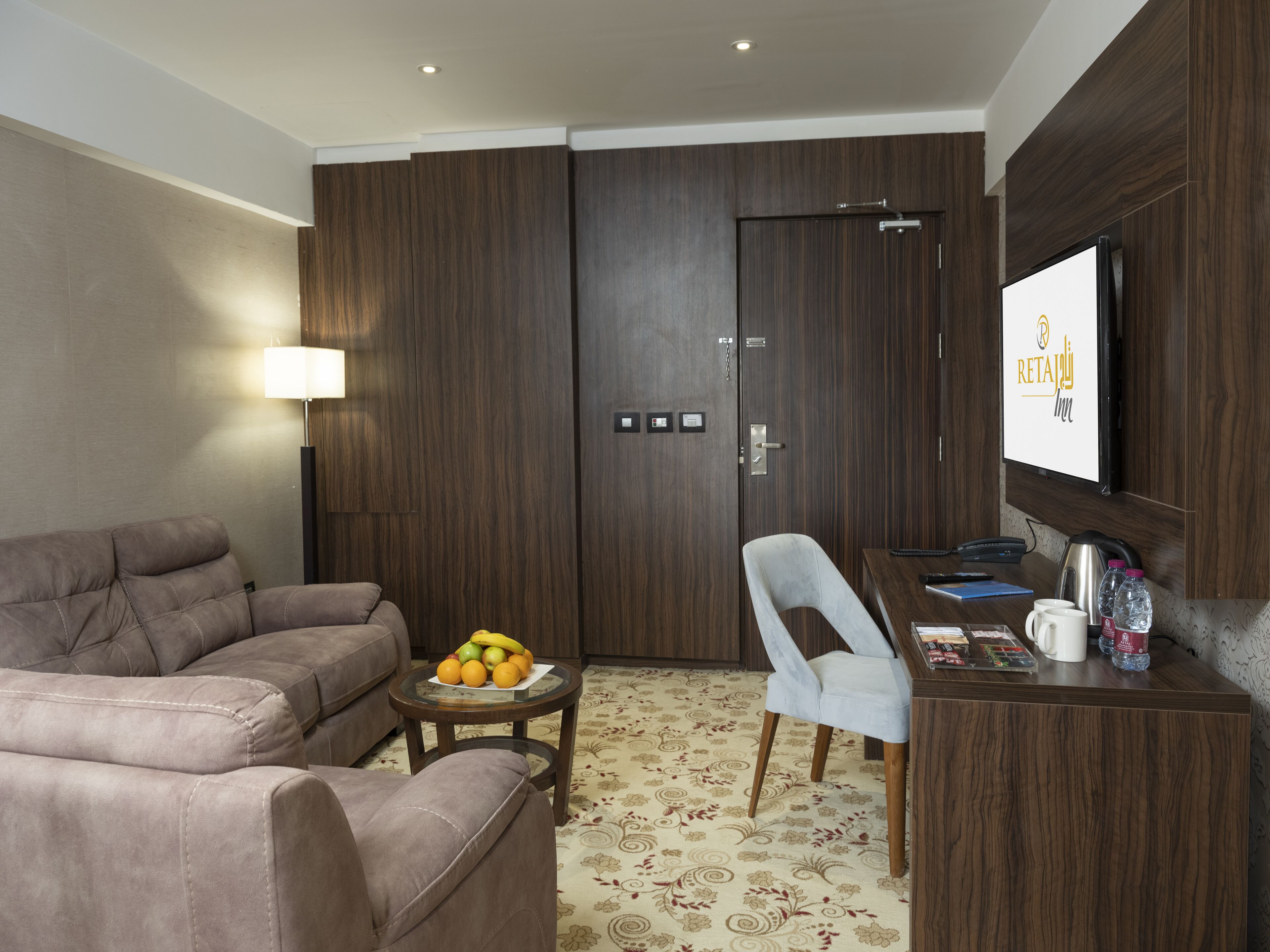 A modern hotel room with a sofa, a dining table with a chair, a fruit bowl, a television, and a minibar.