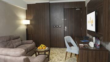 A modern hotel room with a sofa, a dining table with a chair, a fruit bowl, a television, and a minibar.