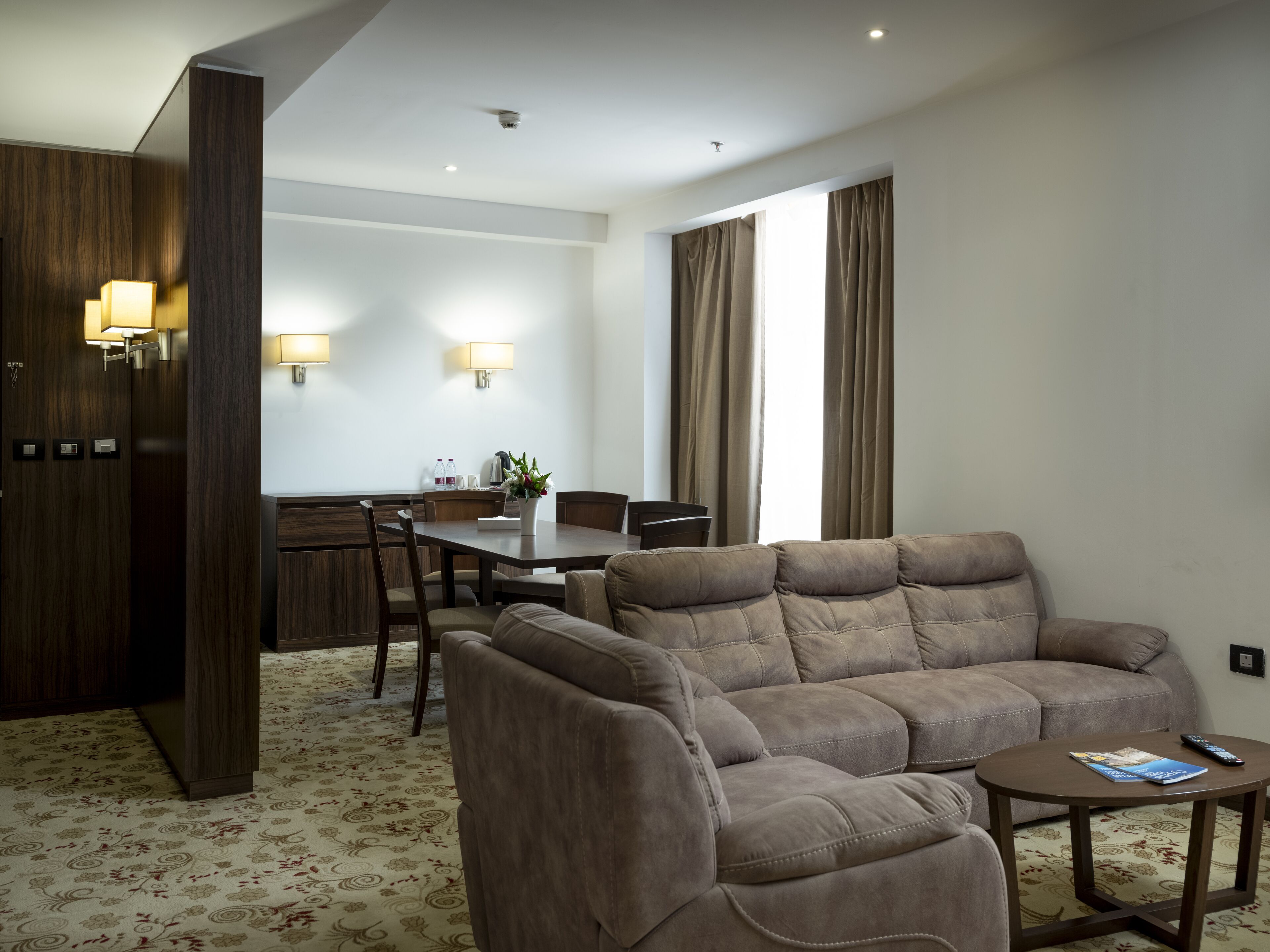 A modern hotel room with a sofa set, a dining table, and a kitchen area.