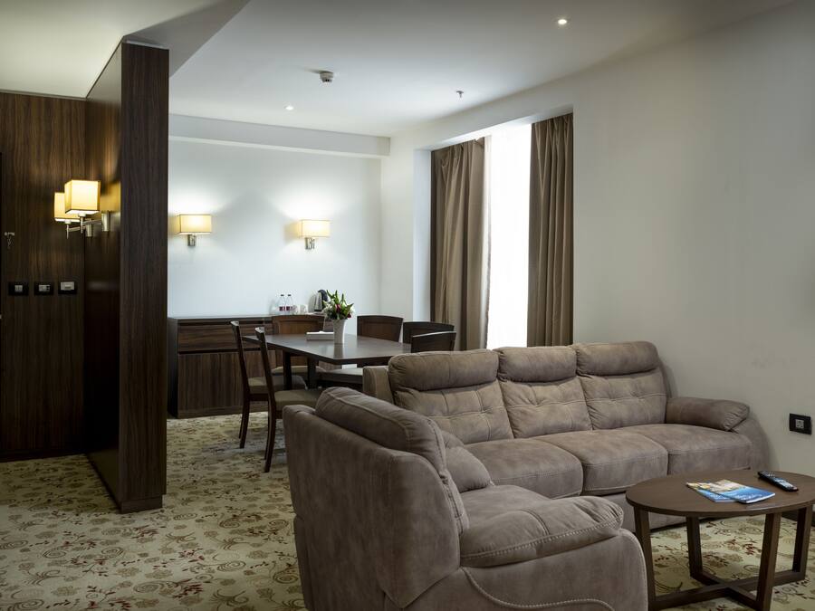 A modern hotel room with a sofa set, a dining table, and a kitchen area.