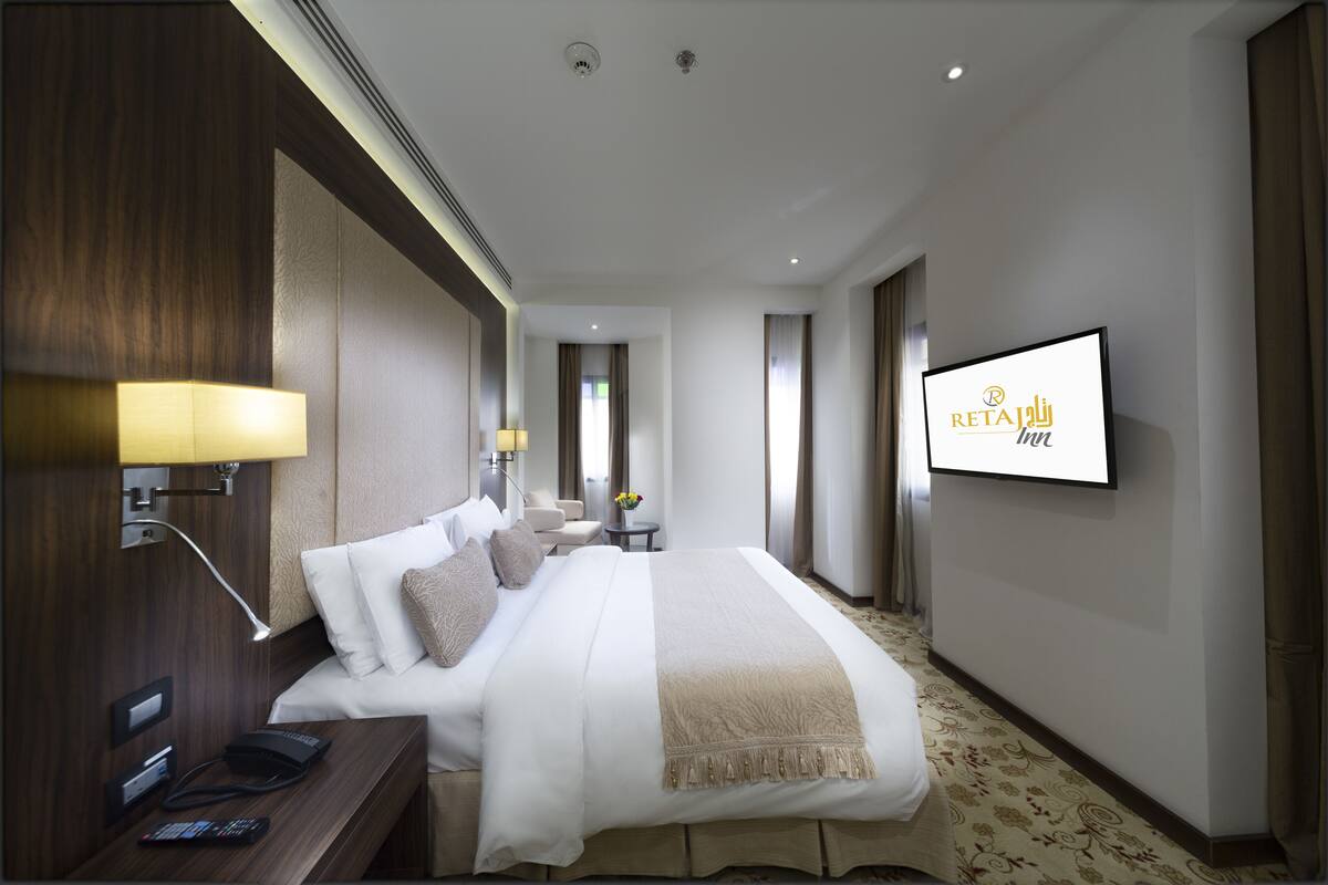 Quarto de hotel com cama, mesa de cabeceira, luminária, TV e vista para o corredor.