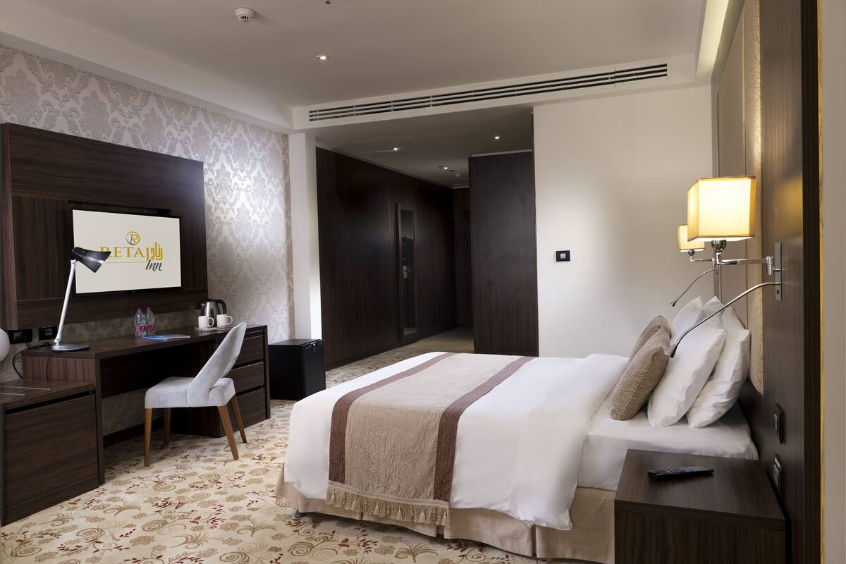 Quarto de hotel com cama, escrivaninha, cadeira e televisão fixada na parede exibindo o nome do hotel.