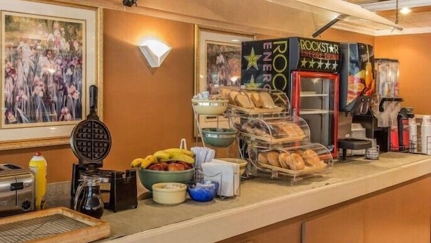 Ein Frühstücksbuffet mit einem Waffeleisen, einem Toaster und verschiedenen Frühstücksangeboten wie Bananen und Brot. An der Wand befinden sich gerahmte Bilder und ein Schild für Rockstar Energy Drink.