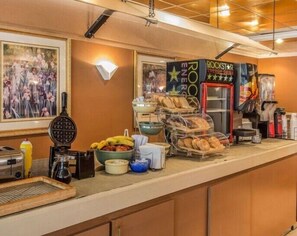 A breakfast bar with a waffle iron, toaster, and various breakfast items like bananas and bread. There are framed pictures on the wall and a Rockstar Energy Drink sign.