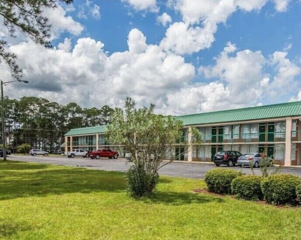 A two-story motel with a green roof, a parking lot with several cars, and a grassy area with a small tree.