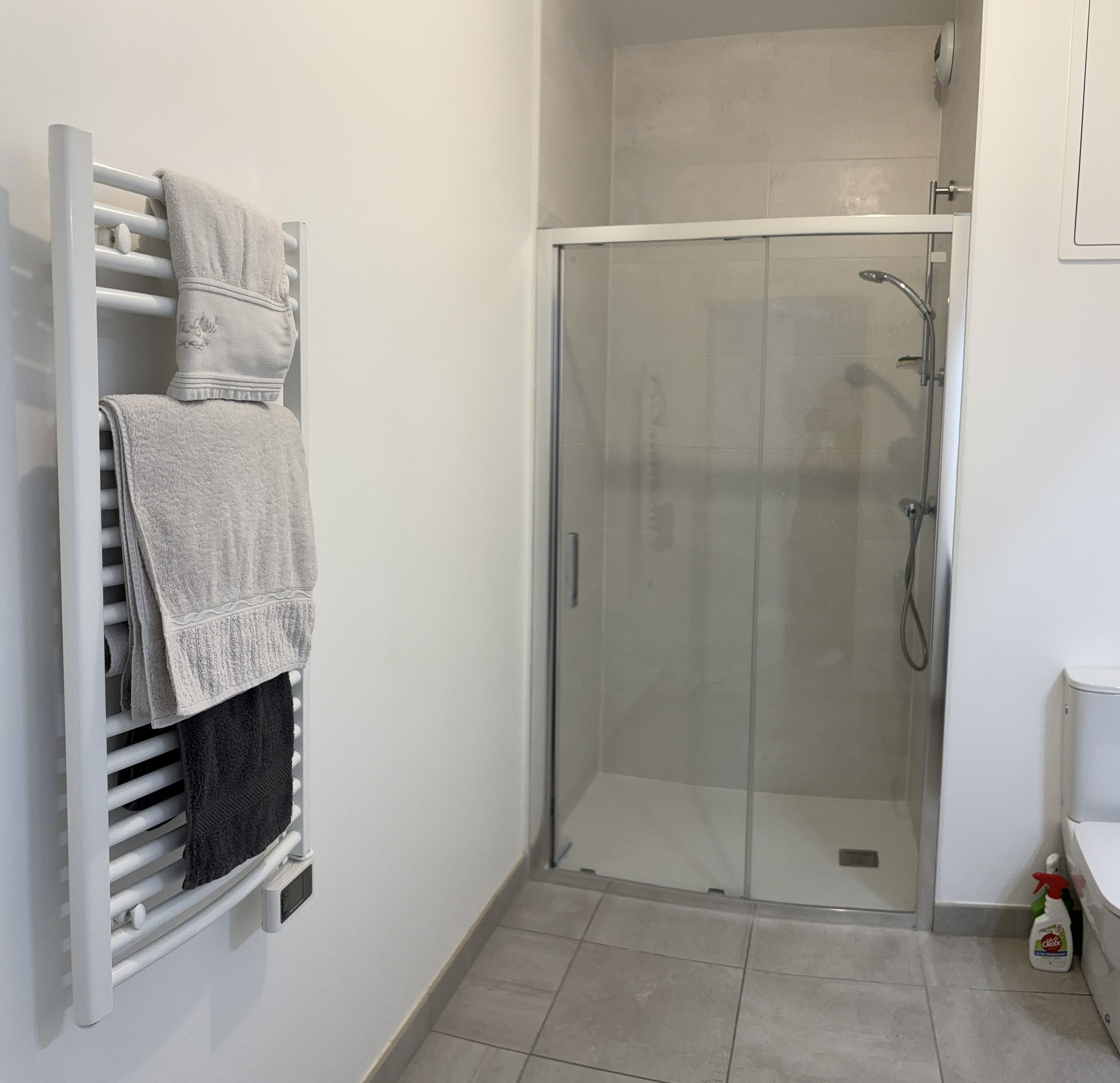 A bathroom with a glass shower enclosure, a towel rack with multiple towels, and a bottle of cleaning product on the floor.
