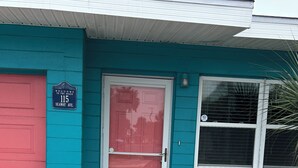 Une maison aux murs bleu sarcelle, avec une porte rouge, deux fauteuils Adirondack turquoise et un panneau indiquant : « Bienvenue à la plage ».