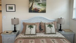 Une chambre à coucher avec un lit, deux tables de chevet, un tableau représentant une scène de plage et une fenêtre avec des stores.