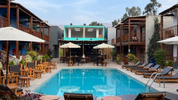 A pool area with lounge chairs and umbrellas, surrounded by buildings with balconies. - VPX HOTEL & Bungalows (Cieneguilla)