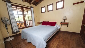 A bedroom with a bed, wooden flooring, a fan, a window with curtains, and a small table with a lamp. - VPX HOTEL & Bungalows (Cieneguilla)