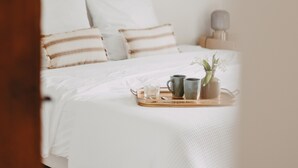 A neatly made bed with white bedding and striped pillows, a bedside table with a lamp, and decorative items on the wall. - West House (Biaudos)