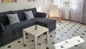 A living room with a dark grey sofa, a small white table, and a chair. There are two paintings on the wall and a window with sheer curtains. - T1 Solaris 200m Praia Wifi (Loulé)