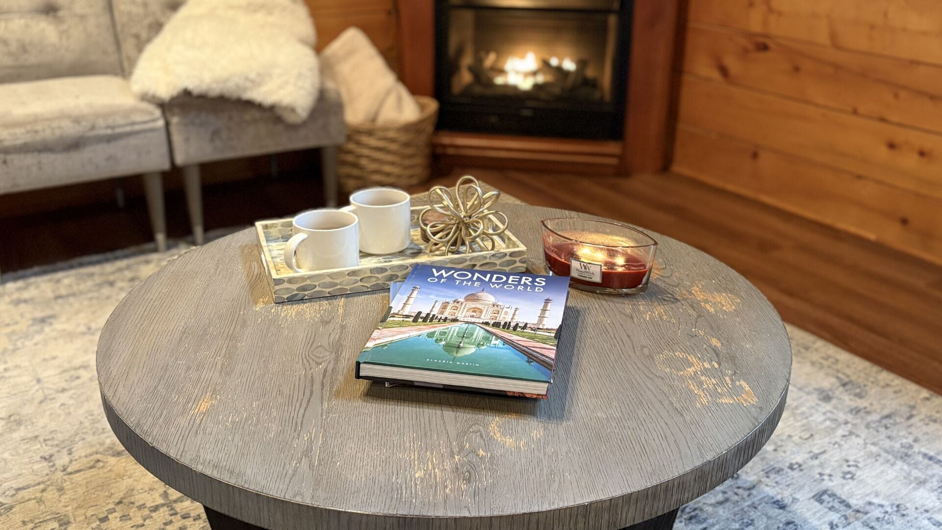 Una mesa redonda de madera con un libro titulado 'Wonders of the World', una vela y una bandeja con tazas y un adorno.