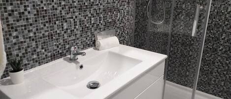 Une salle de bain avec un lavabo blanc, une petite plante en pot, un porte-rouleau de papier toilette et un espace douche doté d’une porte en verre.