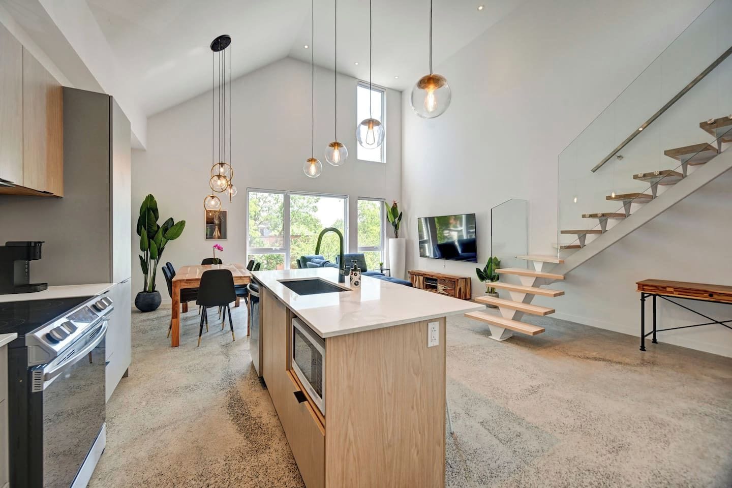 A modern kitchen with a central island, a dining area with a table and chairs, a staircase with a minimalist design, and a large window providing natural light.
