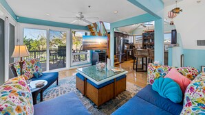 A spacious living room with a glass-top coffee table, a sofa with colorful floral cushions, and a view of the outdoor patio area. - The Salty Conch - Luxury Just Steps To The Beach with Private Pool;2 Golf Carts (Captiva)