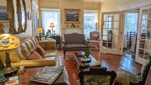 A spacious living room with a leather sofa, a patterned armchair, and a coffee table. A ceiling fan is visible, and there are framed pictures on the walls. - Old Powder House Inn (St. Augustine)