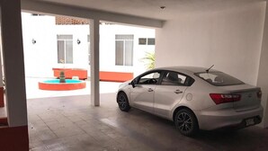 Ein weißes Auto steht auf einem überdachten Parkplatz mit Blick auf ein Gebäude mit orangen Akzenten und einem Springbrunnen im Hintergrund.