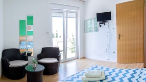 Une chambre d’hôtel avec un lit, deux fauteuils en osier, une petite table, un téléviseur et une porte-fenêtre coulissante donnant sur l’extérieur.